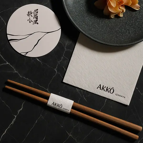 Peças de identidade visual criadas para o restaurante AKKŌ IZAKAYA: hashi com cinta personalizada, guardanapo com logo e descanso de copo com arte japonesa, sobre superfície escura.