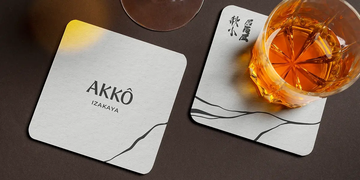 Design de descansos de copo para AKKŌ IZAKAYA: dois modelos quadrados em papel texturizado cinza claro, um exibindo o logo 'AKKŌ IZAKAYA' e outro com caligrafia japonesa, ambos com uma ilustração minimalista preta na base. Um copo com bebida âmbar repousa sobre o segundo.